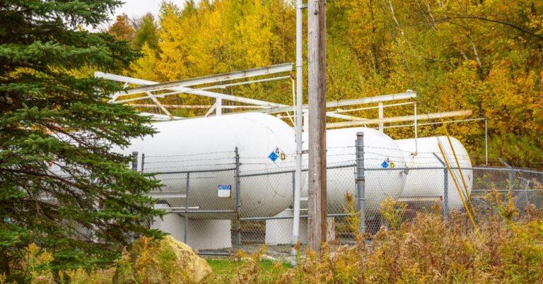 Multiple white propane storage tanks sit behind a chain link fence and different trees, shrubbery, and a tall wooden pole.