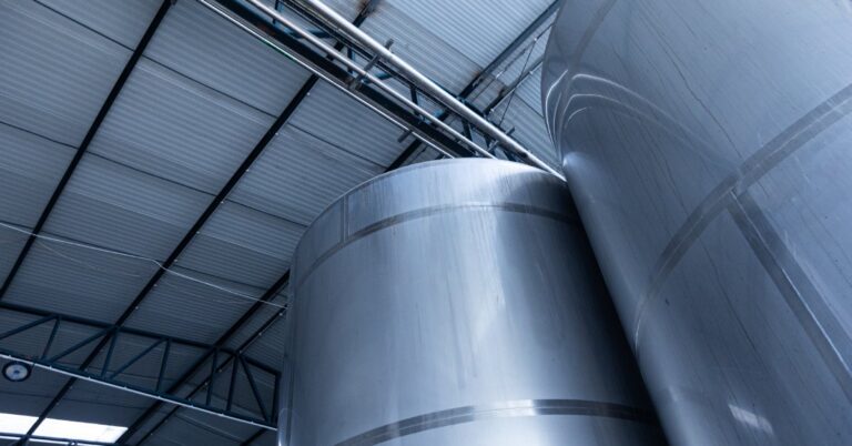 A close-up view of large stainless steel industrial tanks. Above the tanks, the production facility has a large ceiling.