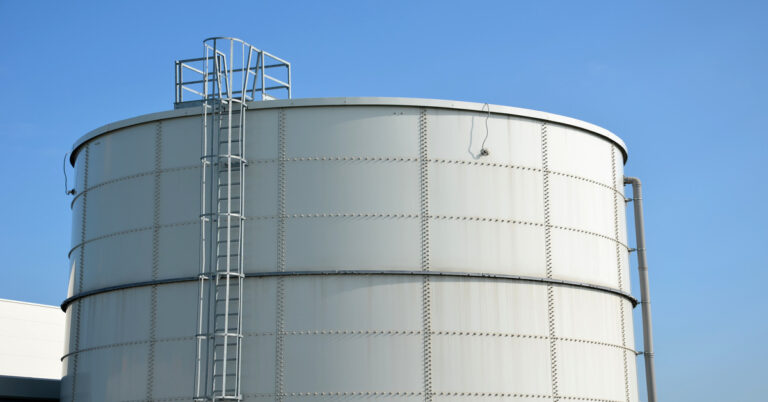 A large white storage tank with a pipe running down it. There is one ladder down the front and another down the side.