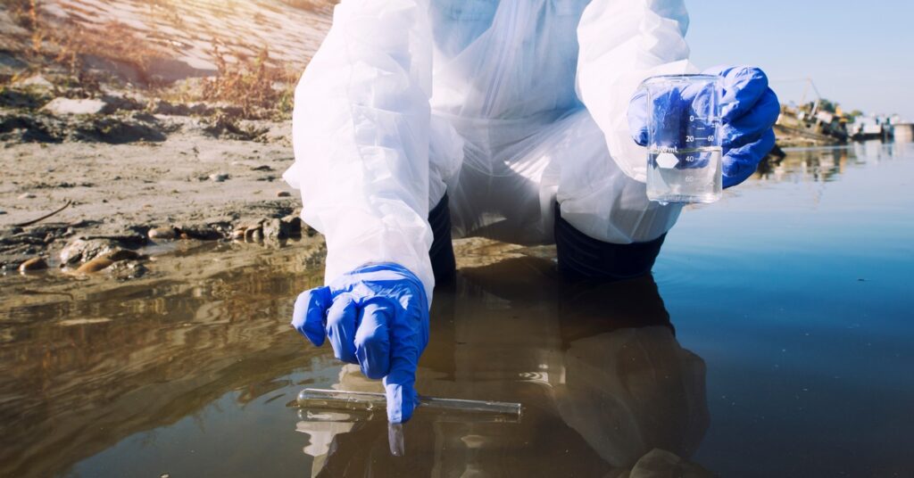A worker wearing PPE crouches down and uses a tool to check the purity of water, holding a beaker in their other hand.