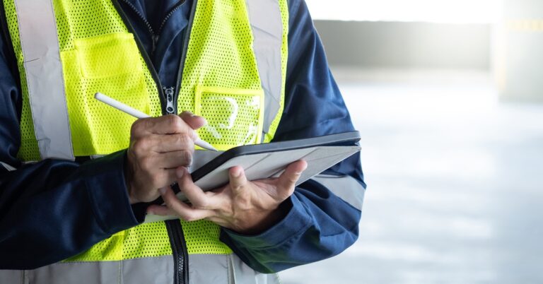 A close up of a worker wearing a high-visibility vest and work clothes types on their tablet with a stylus.