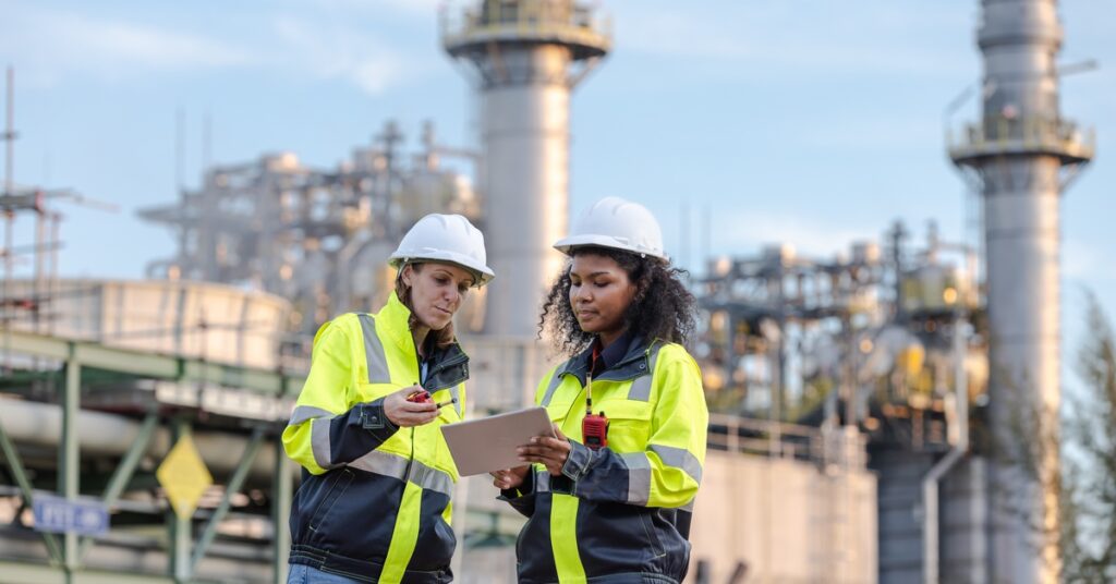 Two workers wearing high-visibility clothes and hard hats look at a clipboard together with an industrial plant behind them.