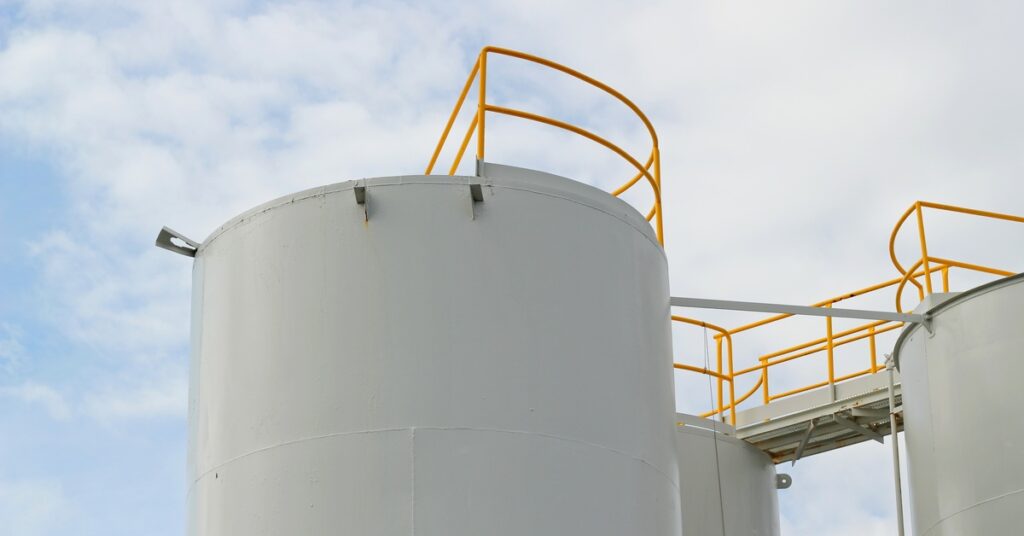 A large storage tank next to some other tanks. The tanks are connected with a small bridge and yellow safety railing.