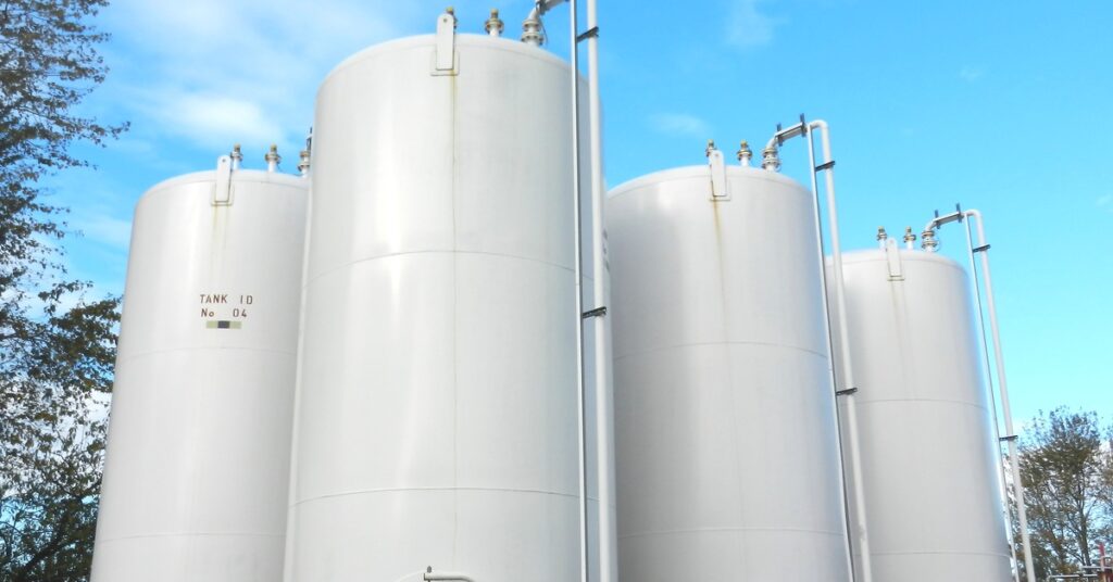 Some tall industrial storage containers standing next to each other with trees and a blue sky and clouds behind it.