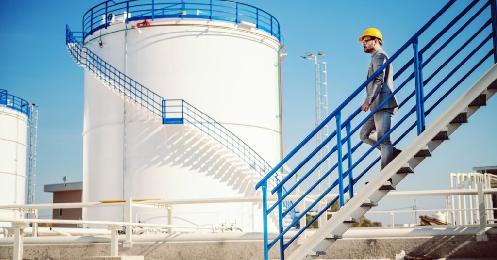 A storage tank is in the distance with a blue staircase going around it. A worker descends a staircase in front of it.