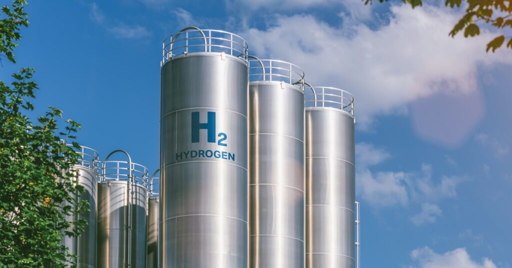 Some large H2 Hydrogen tanks standing next to each other against a blue sky. One tank is labeled "H2 HYDROGEN".