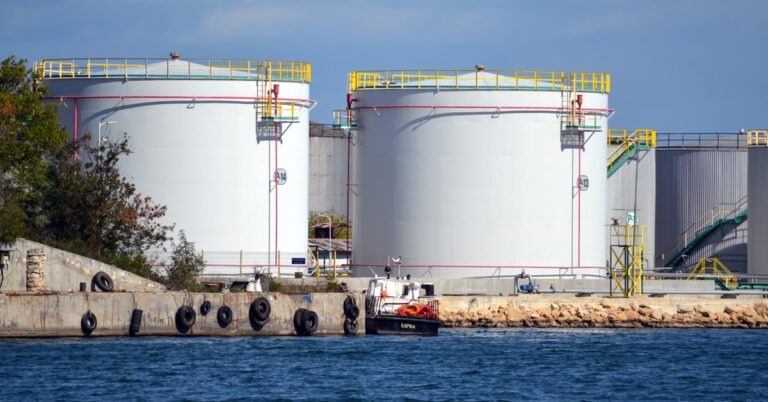 A view across the river from large industrial storage tanks sitting next to each other. The tanks have yellow railings around the top of them.