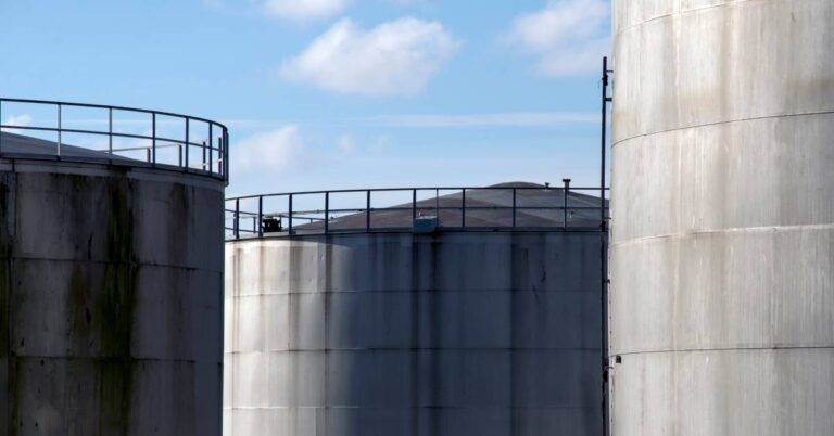Three large industrial storage tanks standing next to each other. Each tank is tall and there is railing at the top.