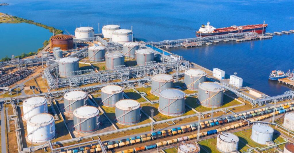 An aerial view of multiple large storage tanks standing next to each other at an industrial facility near the water with boats coming in.