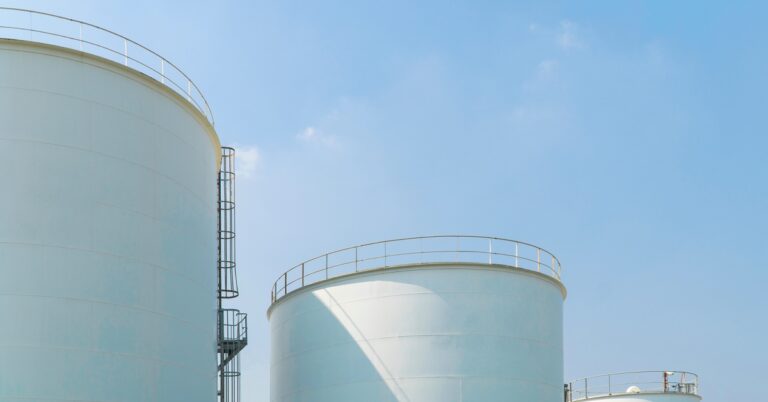 Multiple large storage tanks standing next to each other. Each tank has a ladder leading up to the top next to railing.