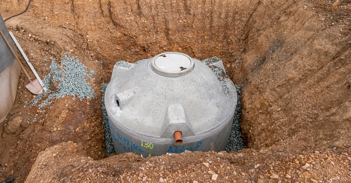 A large gray water cistern sitting in a hole surrounded by sloping dirt. There's a shovel sitting near left side of the hole.