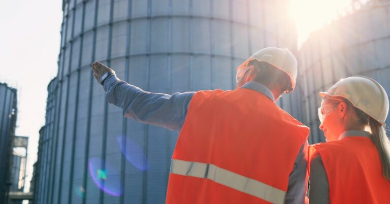 Two workers in hard hats and high-visibility vests supervising a worksite and pointing at large cisterns.