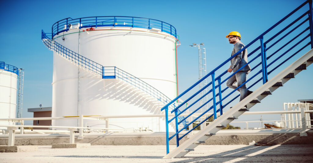 A large storage tank standing in the distance while a businessman walks down a staircase in front of it.
