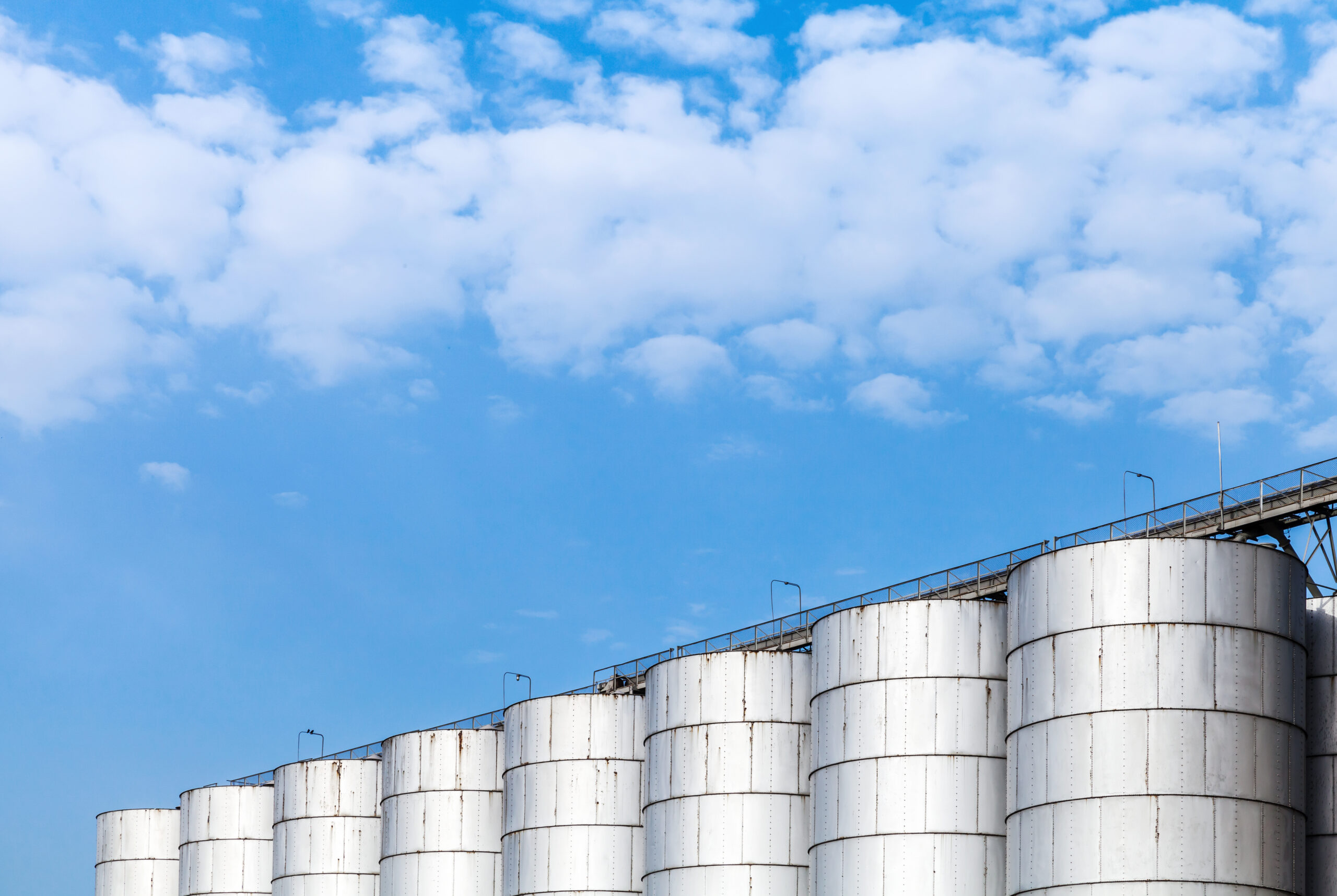 Multiple shining metal storage tanks standing next to each other with a walkway connecting them all. There's a blue sky with clouds in the background.