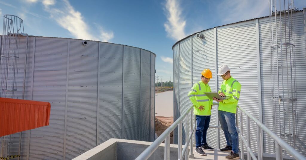 Two men wearing safety jackets and hard hats stand in front of two large industrial tanks, looking at a laptop.