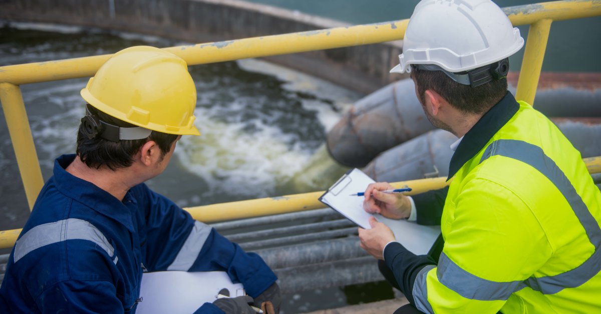 An over-the-shoulder view of a water tank being filled as two men wearing safety jackets and hard hats take notes.