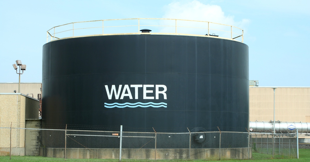 A wide shot of a black water tank that reads "water" on the side with a wave motif. The tank and building sit against a blue sky.