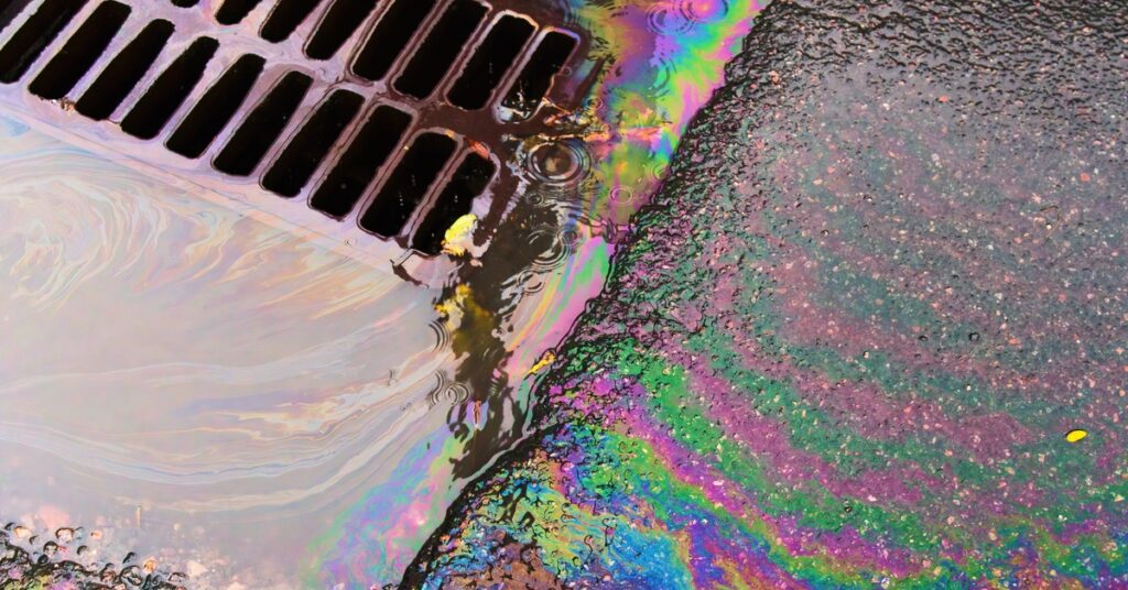 A large oil slick covering an asphalt road. The oil is spilling into a small metal grate on the ground.