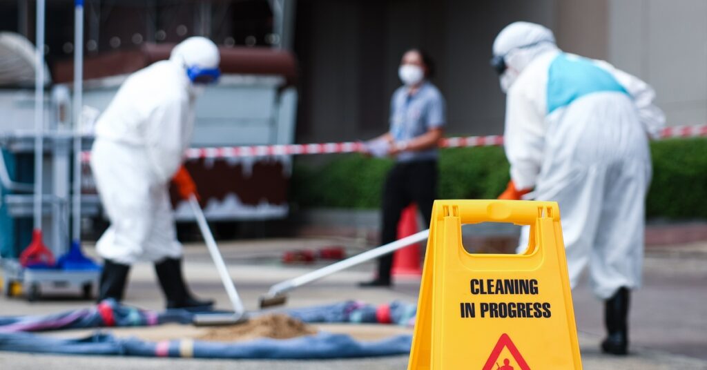 Two workers cleaning a spill while another worker stands nearby. A sign close to the spill reads “Cleaning in Progress.”
