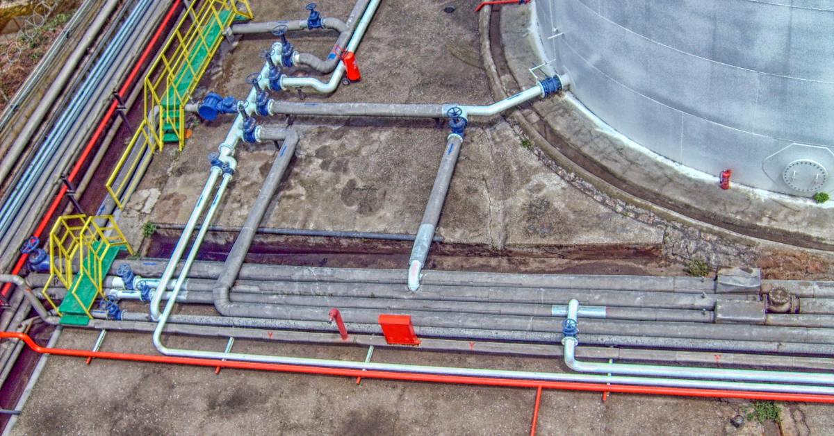A focus on the pipelines leading from the base of a storage tank, leading to walkways and pumps of various colors.