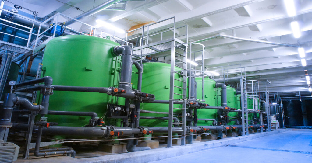 The inside of an industrial plant with six or more green water treatment tanks lined up in a row with various components attached.