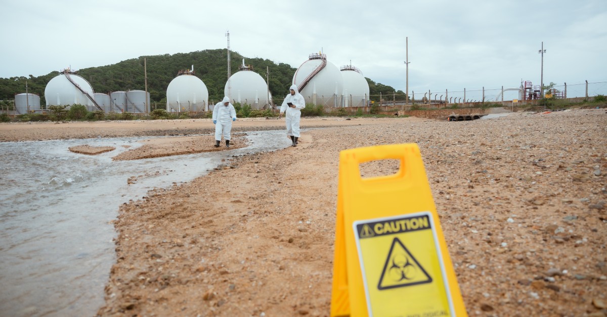 A chemical storage facility near a beach with two workers in protective suits walking the beach near a Caution sign.