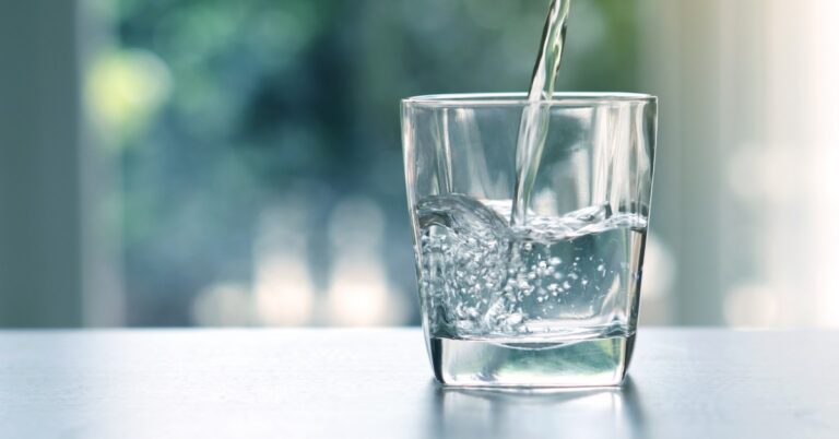A close-up of a clear glass with clean water being poured into it from above with a blurry window in the background.