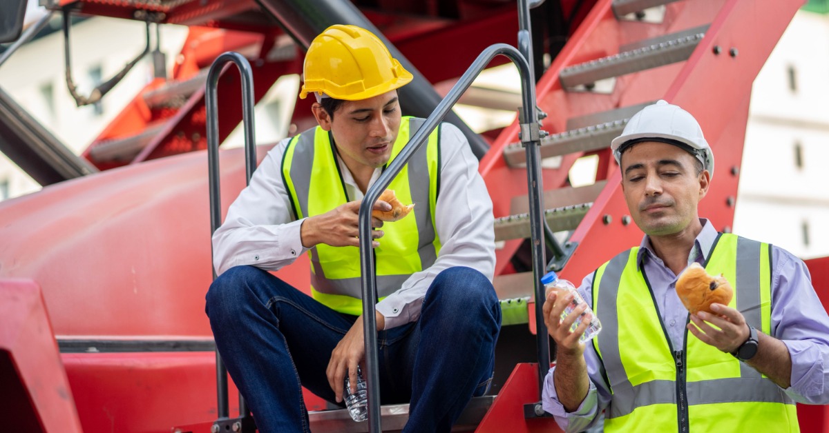 Two workers wearing hard hats and safety vests resting and eating outdoors on a lunch break at a facility.