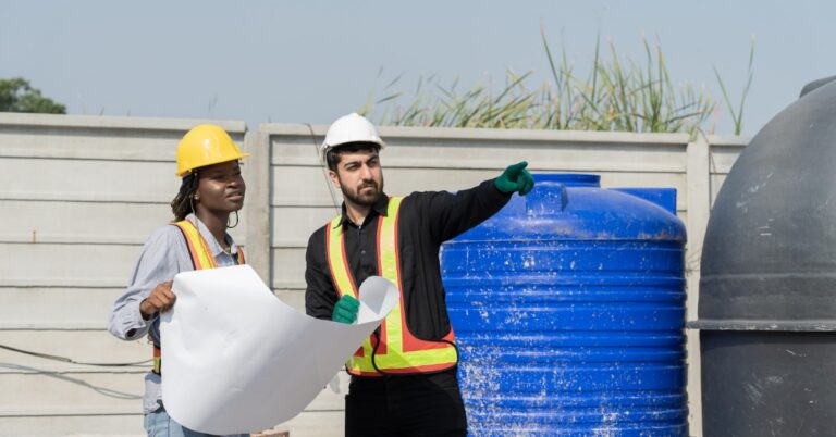 A man and a woman wearing hard hats and safety vests inspect a blueprint while pointing at something out of view.