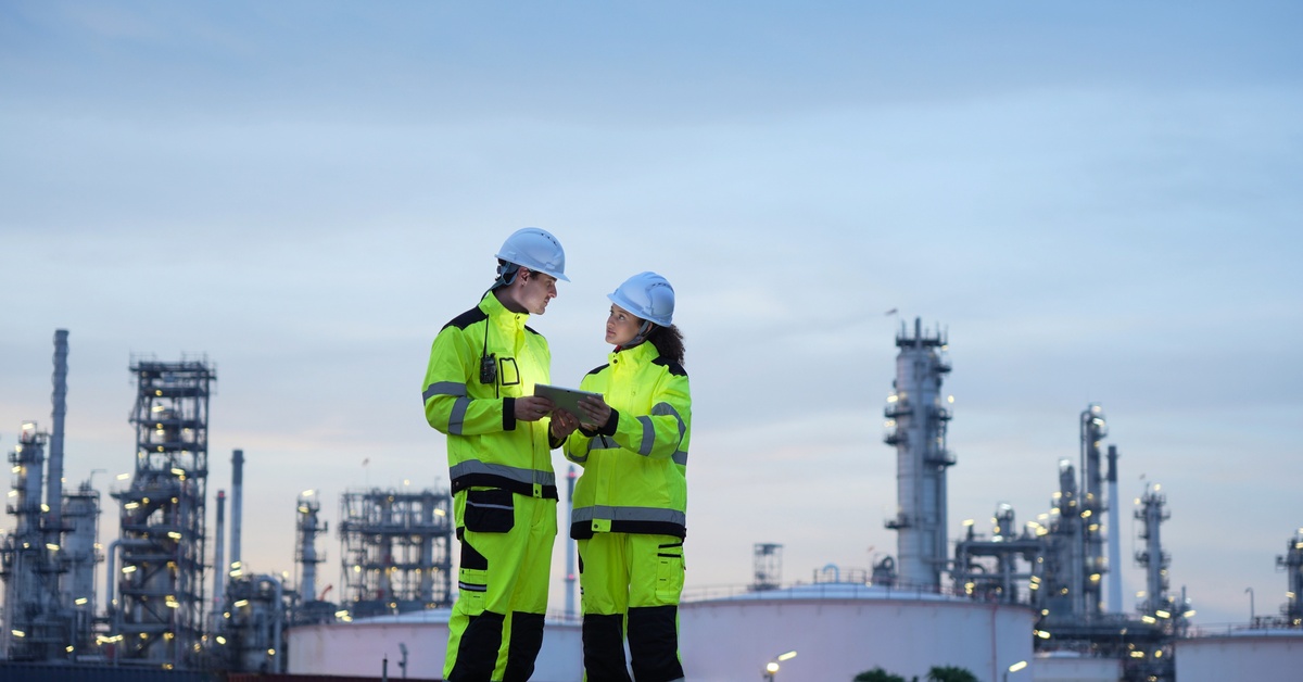 Two people wearing green safety suits and white hard hats examining a tablet at a manufacturing facility.