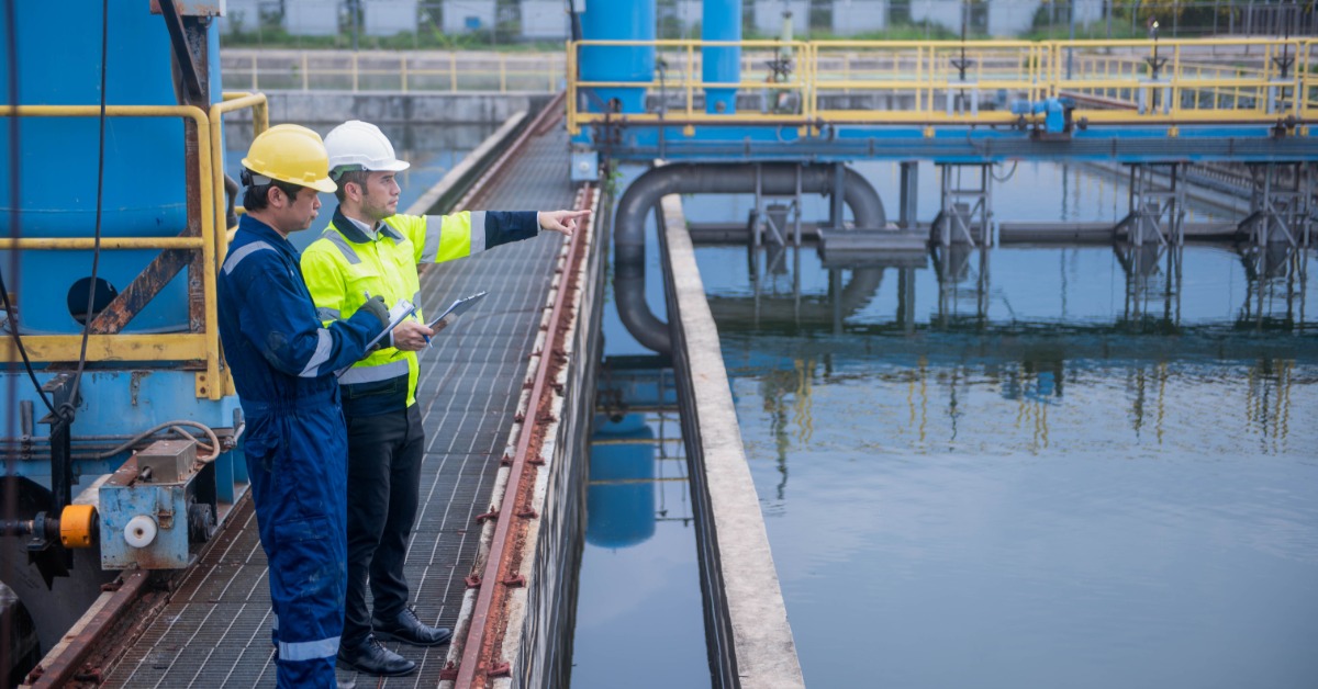 Two men wearing protective hard hats and safety jackets observing a water treatment plant reservoir.