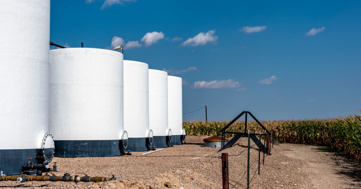A lineup of four secondary water containers on a farm property, near rows of cornfields and a blue sky background.