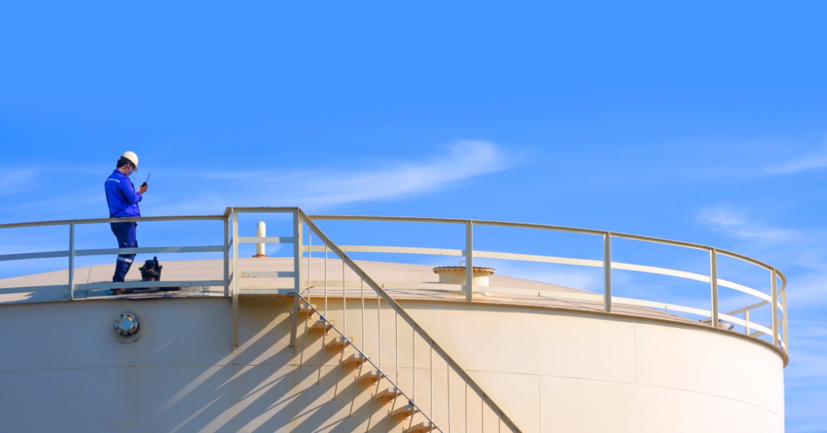 A worker wearing a hard hat and safety vest using a radio while standing on a metal tank platform under a blue sky.