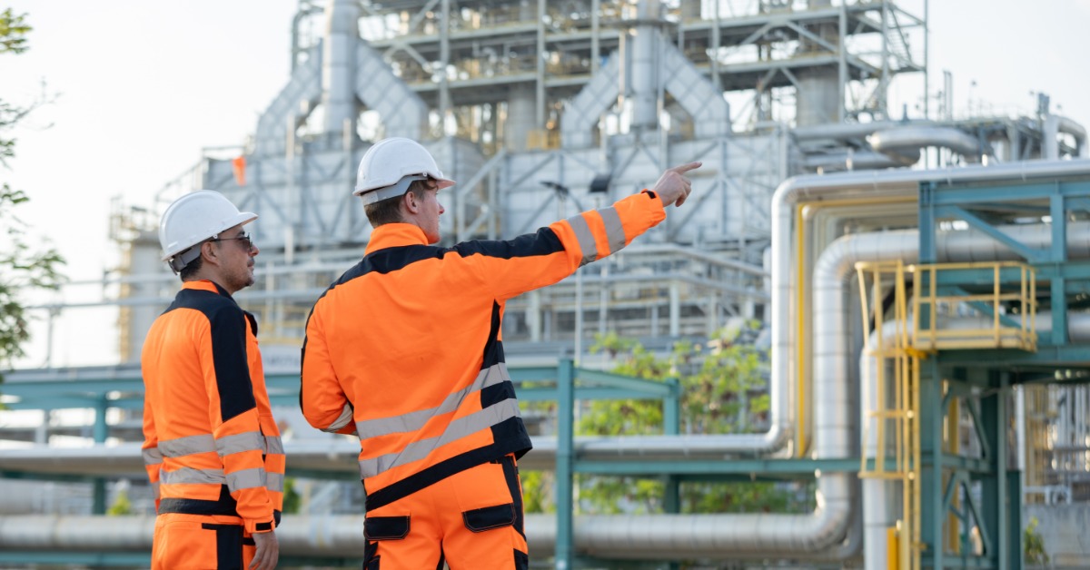 Two workers in orange safety gear talking near metal pipelines and industrial structures at an outdoor plant.