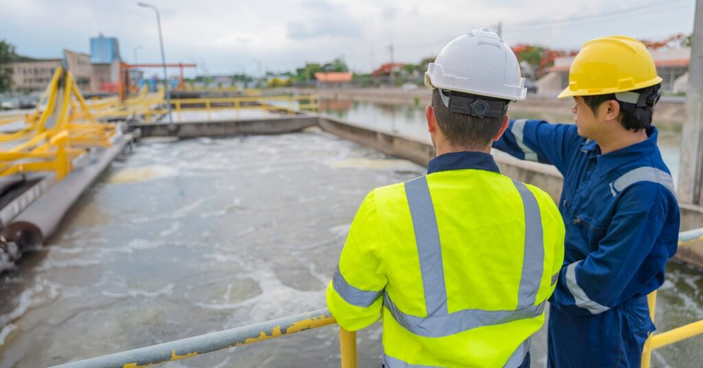 two people in hard hats reviewing a clipboard on a metal walkway at an outdoor water treatment plant.