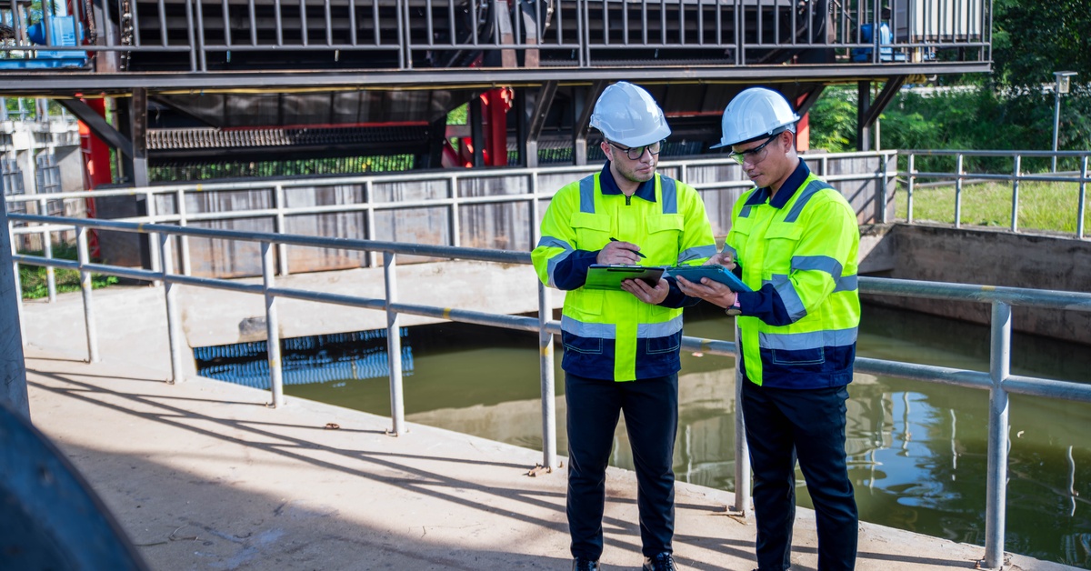 two people in hard hats standing beside industrial water filter equipment and reviewing documents outdoors.