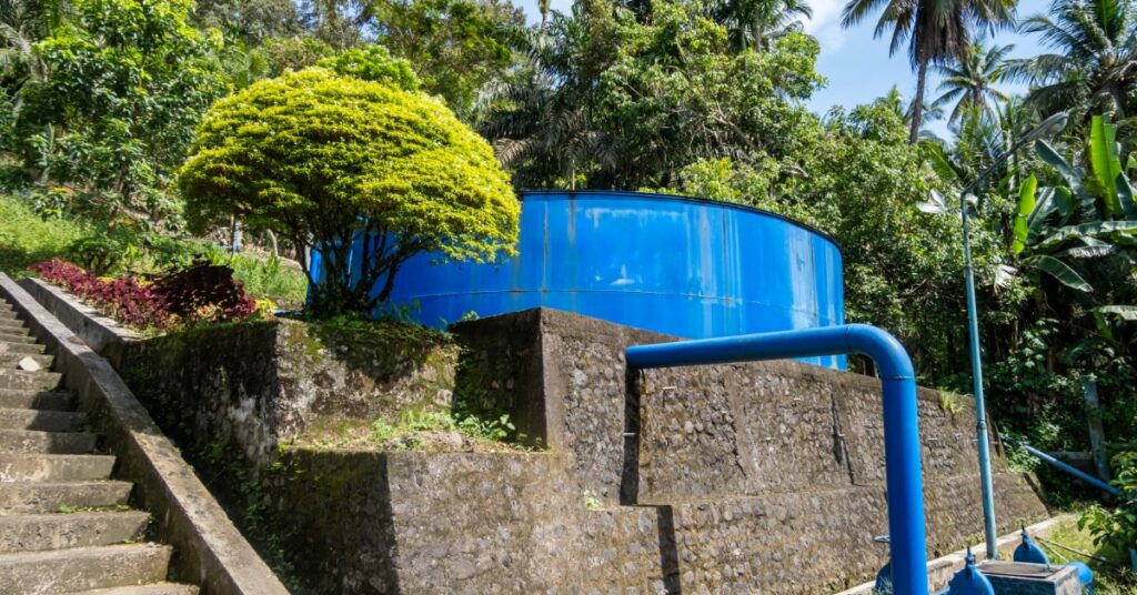 a blue storage tank and large pipe beside stone walls and steps, surrounded by dense green trees and palms.