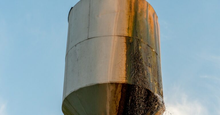 a rust-stained metal water tank leaking water down one side against a blue sky with light clouds behind it.
