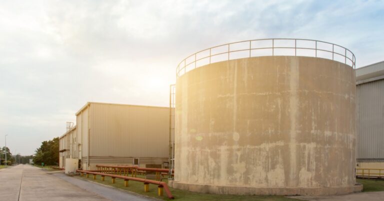 A large concrete tank with a top railing beside industrial buildings, red pipes, grass, and pavement under a cloudy sky.