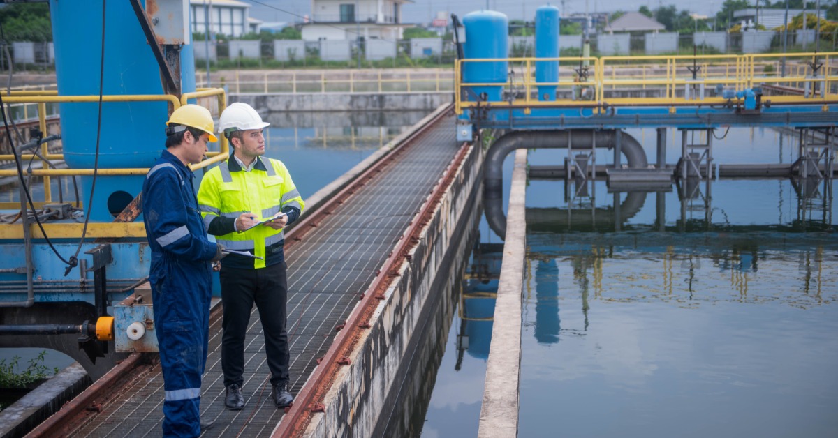 Two people wearing hard hats reviewing paperwork beside pipes, tanks, gauges, and equipment in a water plant.