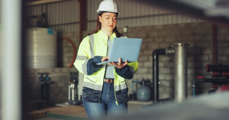A woman wearing a hard hat typing on a laptop beside pipes, tanks, and gauges inside a water facility.