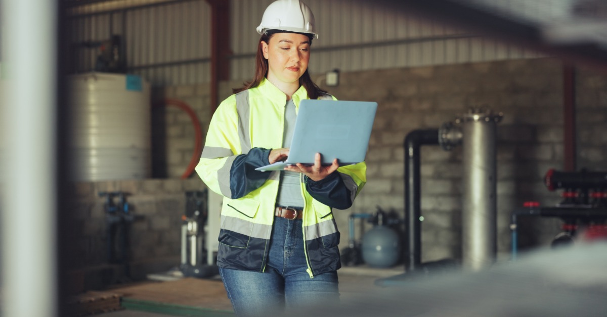 A woman wearing a hard hat typing on a laptop beside pipes, tanks, and gauges inside a water facility.