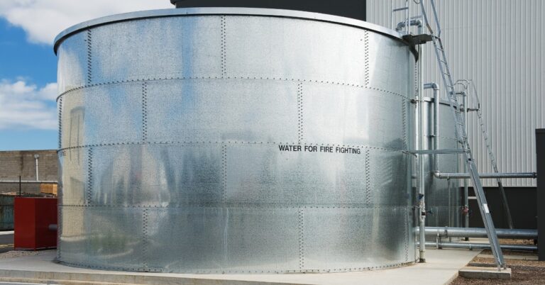 A large silver metal water tank beside an industrial building, with pipes, ladders, and parking spaces nearby.