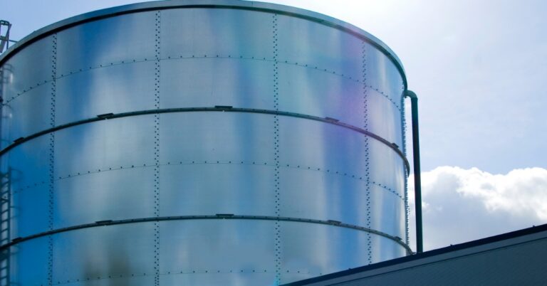 Bright sun glare hangs above a curved metal water tank with riveted panels beside a low building and pale blue sky.