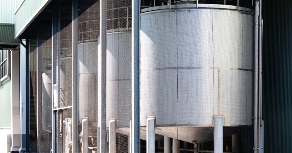Mesh panels enclose metal water tanks outside a green metal building, with pipes and white posts nearby.
