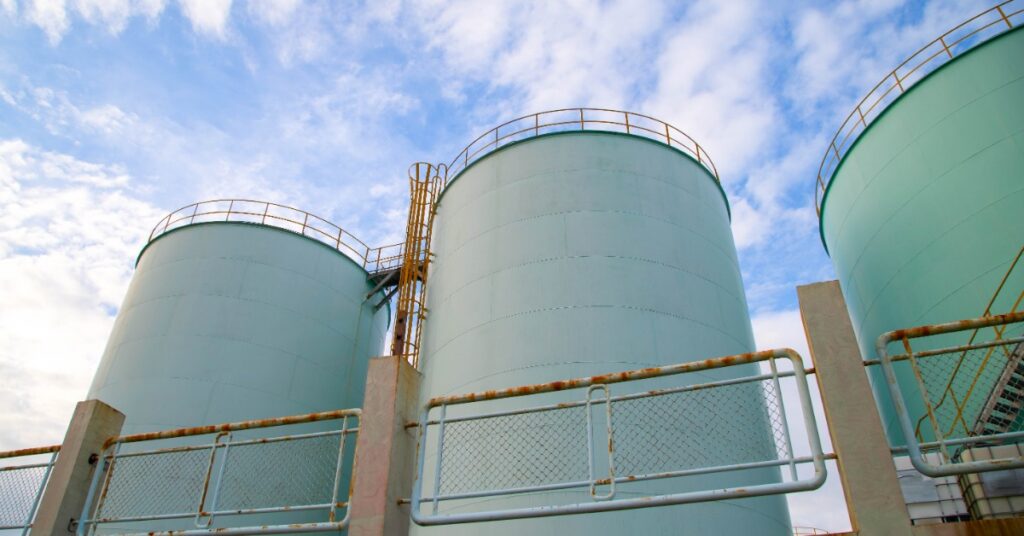 Large pale green storage tanks standing behind a chain-link fence beneath a bright sky with scattered clouds.