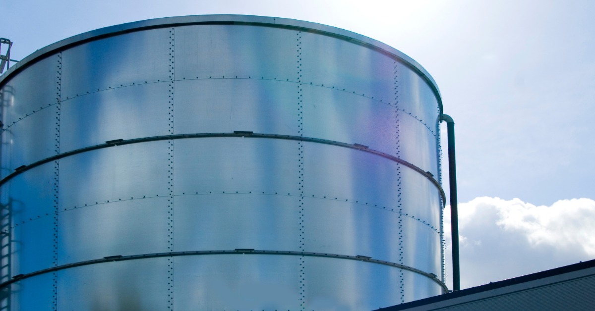 A large cylindrical silver metal water storage tank with horizontal seams and a vertical pipe beneath a cloudy sky.