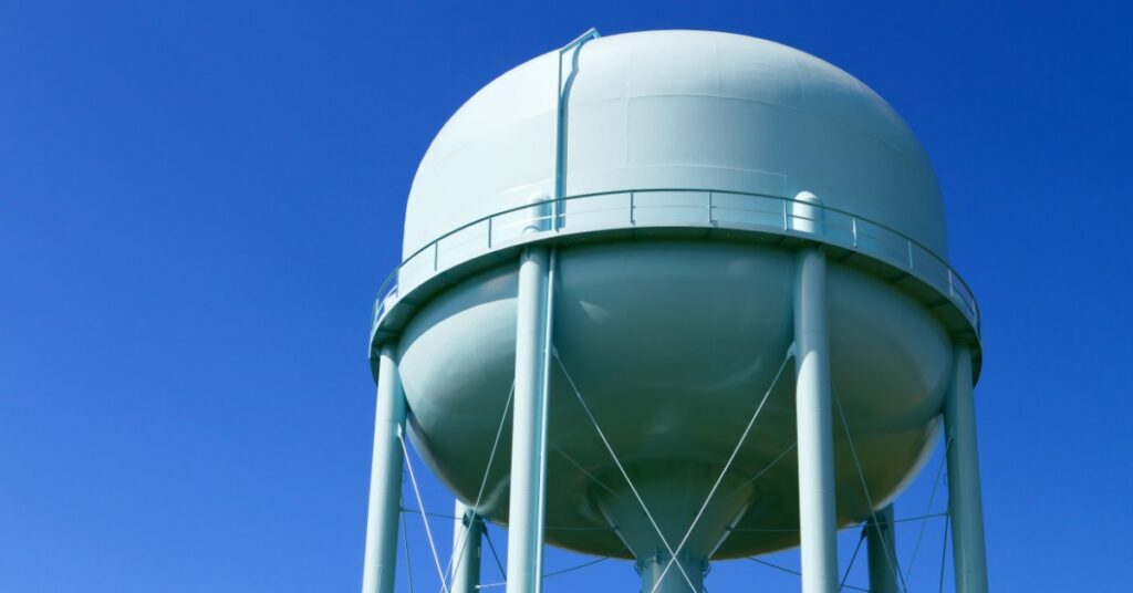 A large pale blue elevated water tower supported by metal columns and cross braces beneath a clear blue sky.